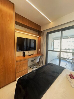 Ett hotellrum med en säng, ett skrivbord, en stol, en TV och en balkong med ett bord och stolar.