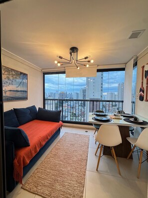 A modern living room with a sofa, dining table, and chairs. A city view is visible through large windows.