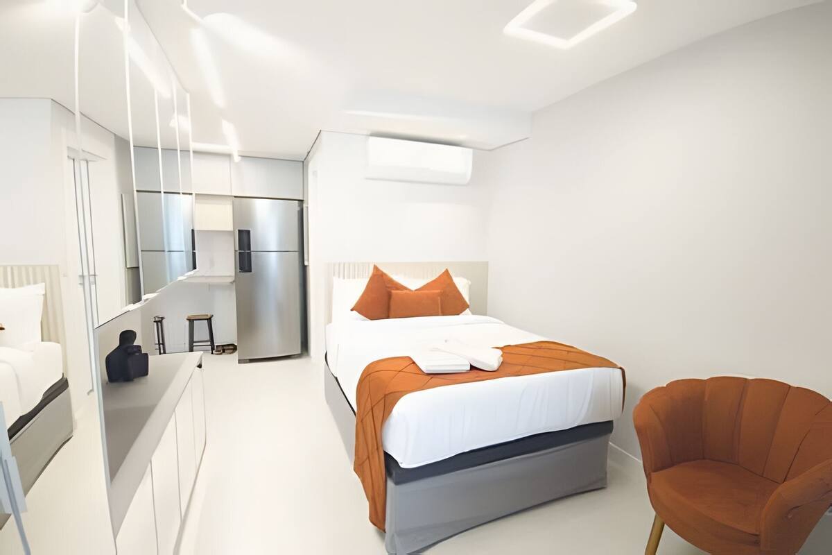 A modern hotel room with a bed, a chair, a wardrobe, a mirror, a refrigerator, and a small table.