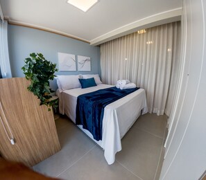 A hotel room with a bed, a plant, artwork on the wall, and a curtain.