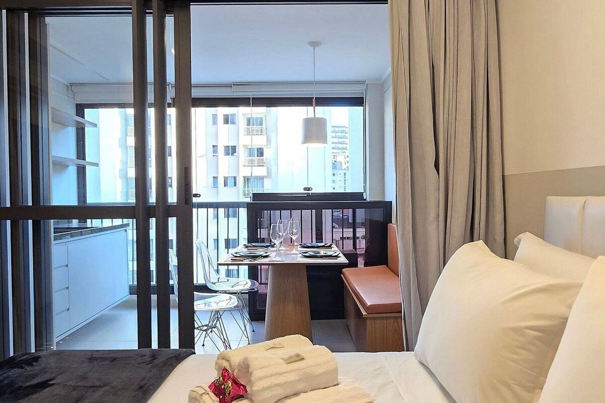 A hotel room with a bed, a small table with chairs, a balcony with a view of buildings, and a large window with curtains.