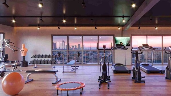A modern gym with various exercise equipment, a large window with a city view, and a wooden floor.