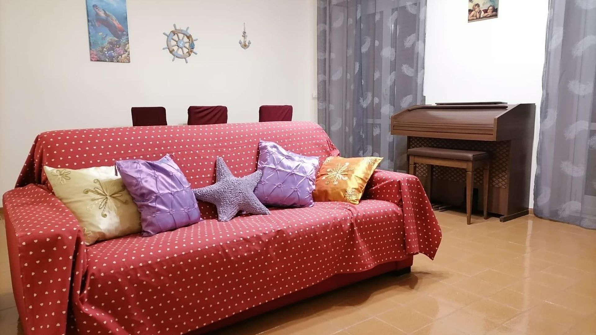 Un canapé rouge à pois et orné de motifs en forme de cœur, agrémenté de plusieurs coussins, d’une table d’appoint en bois et d’une décoration murale représentant un gouvernail de navire.