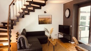 Uma sala de estar moderna com um sofá preto, escada de madeira, televisão de tela plana e uma janela grande.