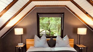 Quarto com teto inclinado, ampla janela com vista para árvores, cama com várias almofadas e duas luminárias de cabeceira.