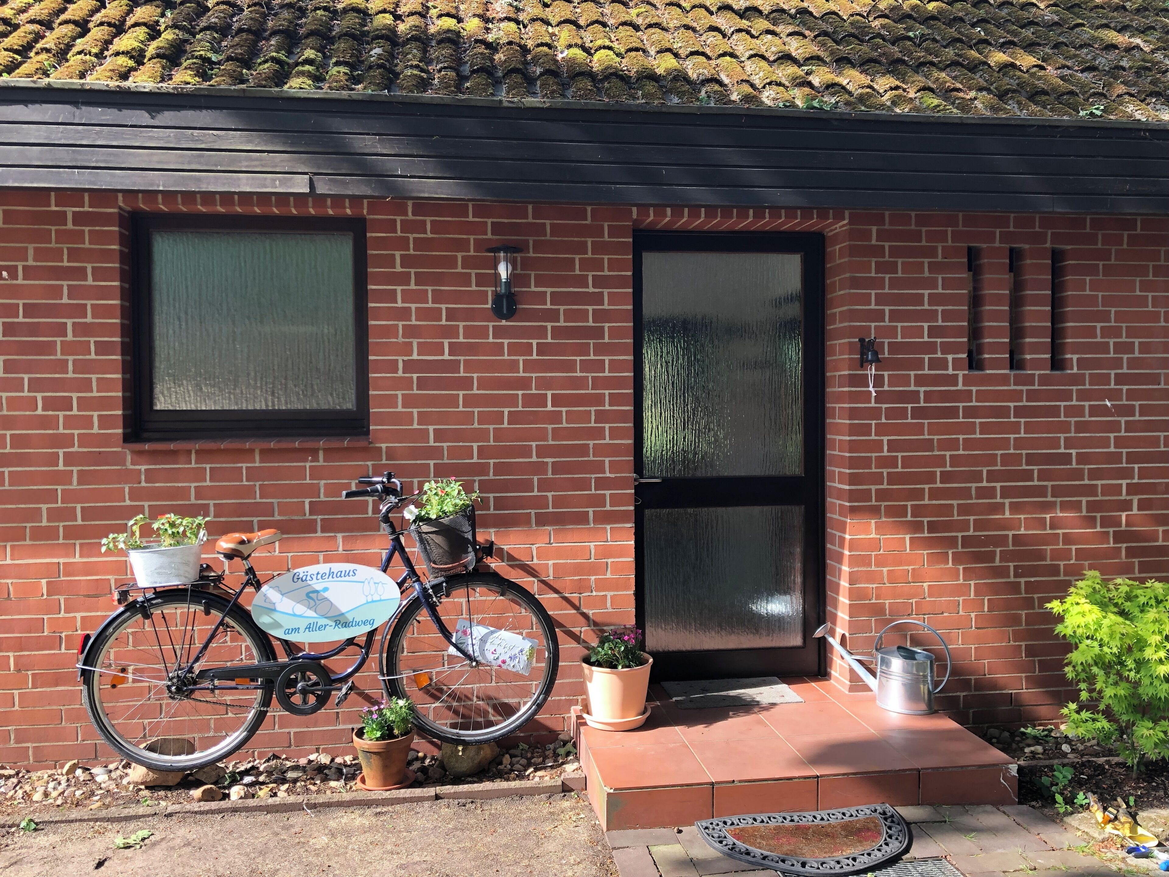 Un bâtiment en briques avec une porte et des fenêtres noires, un vélo avec un panier et des plantes en pot.