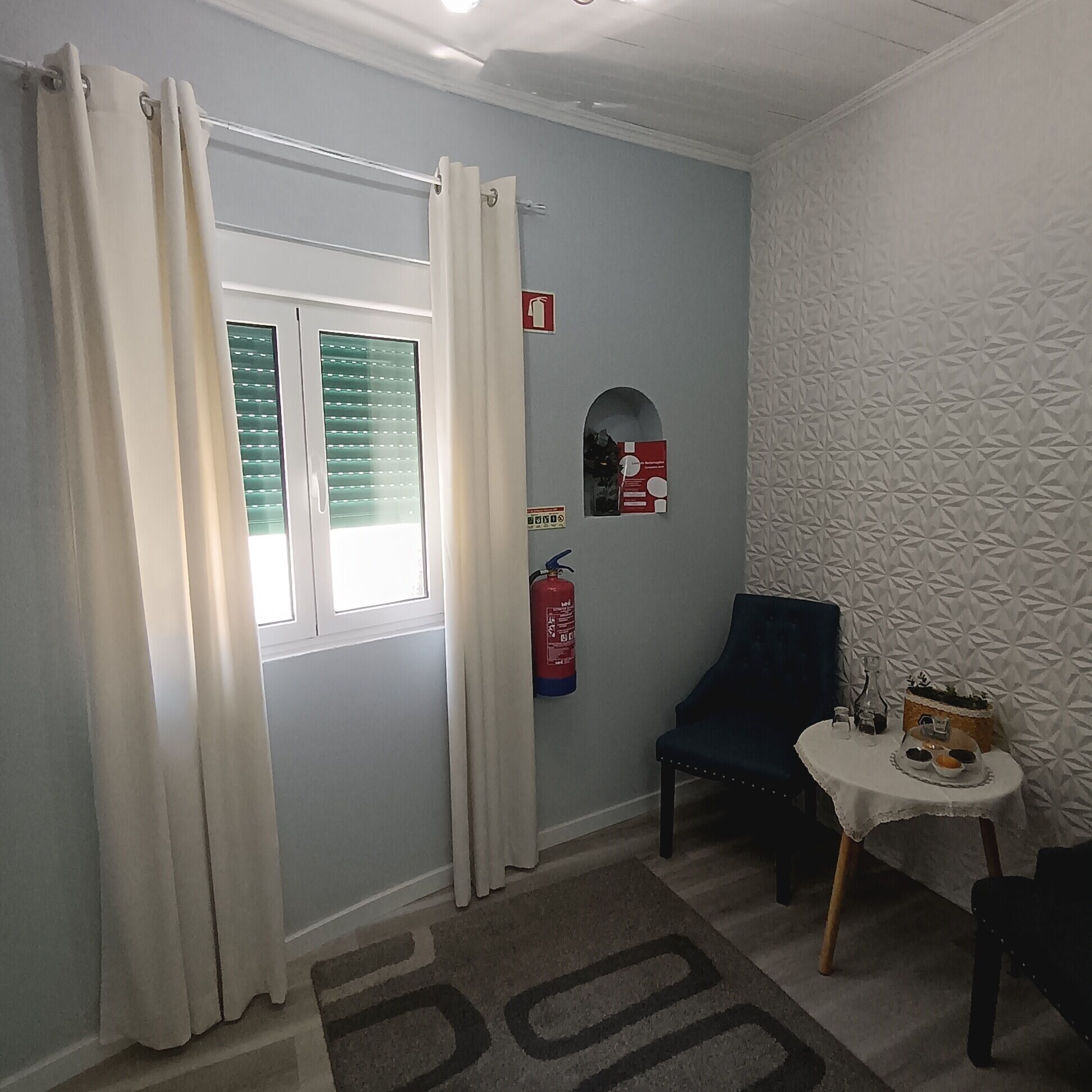 Une pièce avec une fenêtre, un extincteur, une chaise et une petite table sur laquelle se trouvent un panier et une bouteille.