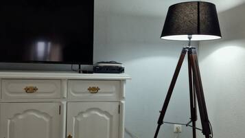 Una stanza con una TV a schermo piatto su un mobile bianco, una lampada da terra e una presa elettrica.