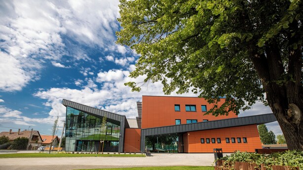 A modern building with a glass facade and a brick exterior, surrounded by a well-maintained lawn and a large tree. - A22 Hotel Gyál (Gyal)