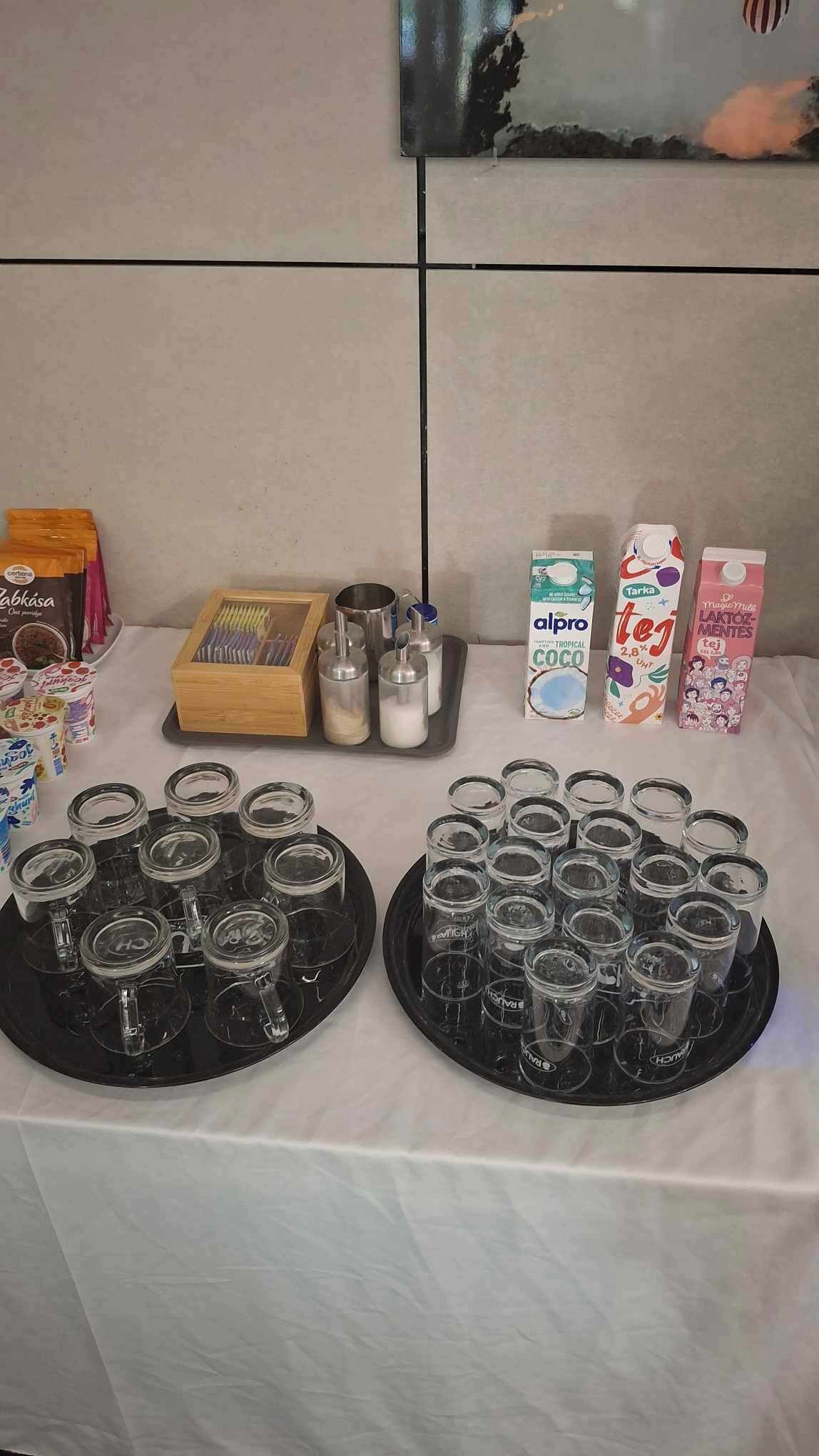 A table with various food items and drinks, including jars, cartons, and a tray of glasses.