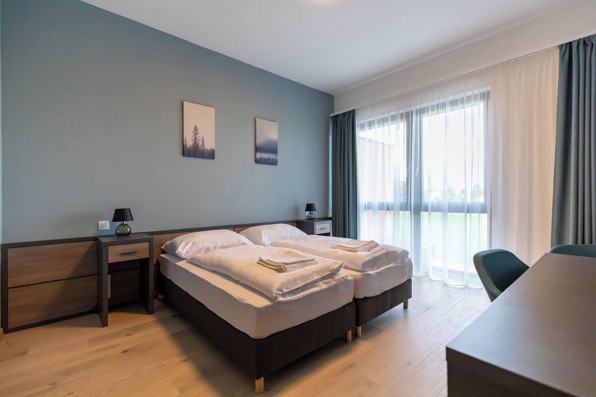 Une chambre d’hôtel moderne dotée d’un grand lit, de tables de chevet, d’un bureau et offrant une vue sur l’extérieur grâce à de grandes fenêtres.
