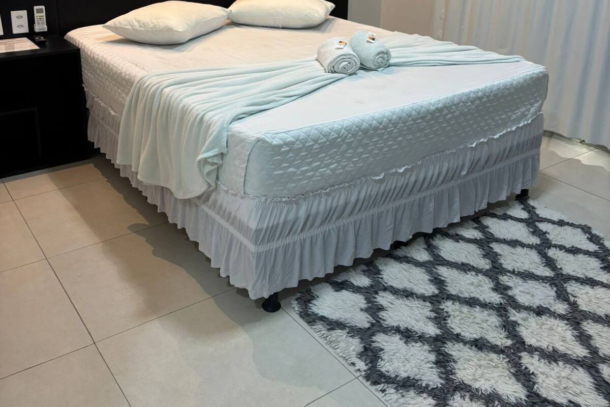 A neatly made bed with white bedding and a decorative pillow, a black headboard, a small bedside table with a phone, and a patterned rug on a tiled floor.