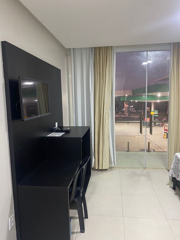 A hotel room with a black desk, a flat-screen TV mounted on the wall, a sliding glass door to the outside, and a view of a gas station at night. - HOTEL QUATRO CORAÇÕES (Arapiraca)