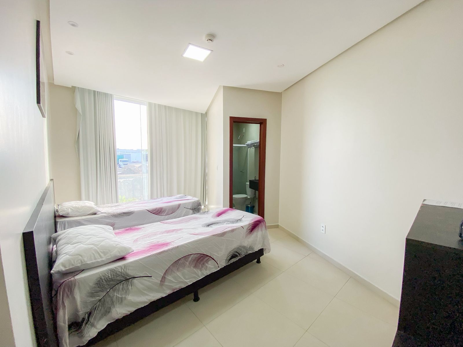 Camera d'albergo con due letti, un'ampia finestra con tende, un bagno parzialmente visibile attraverso una porta e un piccolo angolo cottura.
