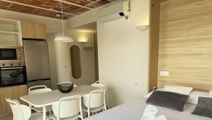 Quarto de hotel moderno com cama, mesa de jantar com cadeiras, área de cozinha com refrigerador e unidade de ar condicionado.