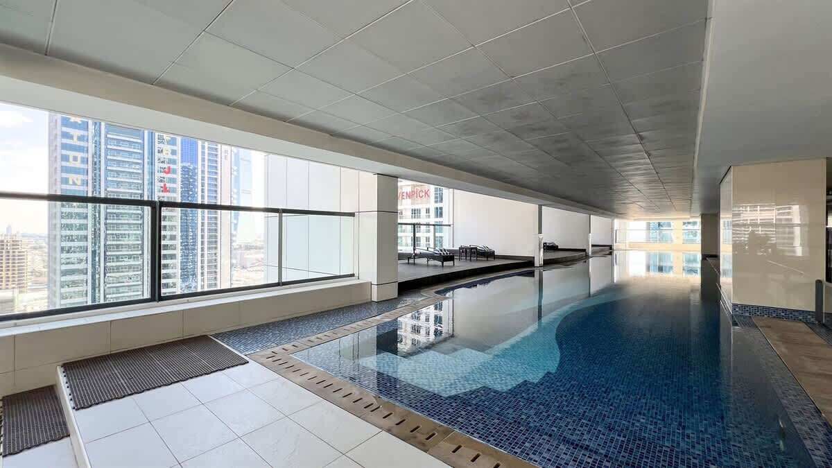 A modern indoor swimming pool with clear glass walls and a tiled floor, surrounded by high-rise buildings.