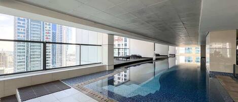A modern indoor swimming pool with clear glass walls and a tiled floor, surrounded by high-rise buildings.