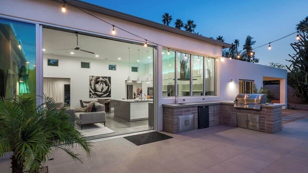 Une maison moderne dotée d’une grande porte vitrée donnant sur un espace intérieur, d’un patio avec un barbecue et d’une vue sur des palmiers.