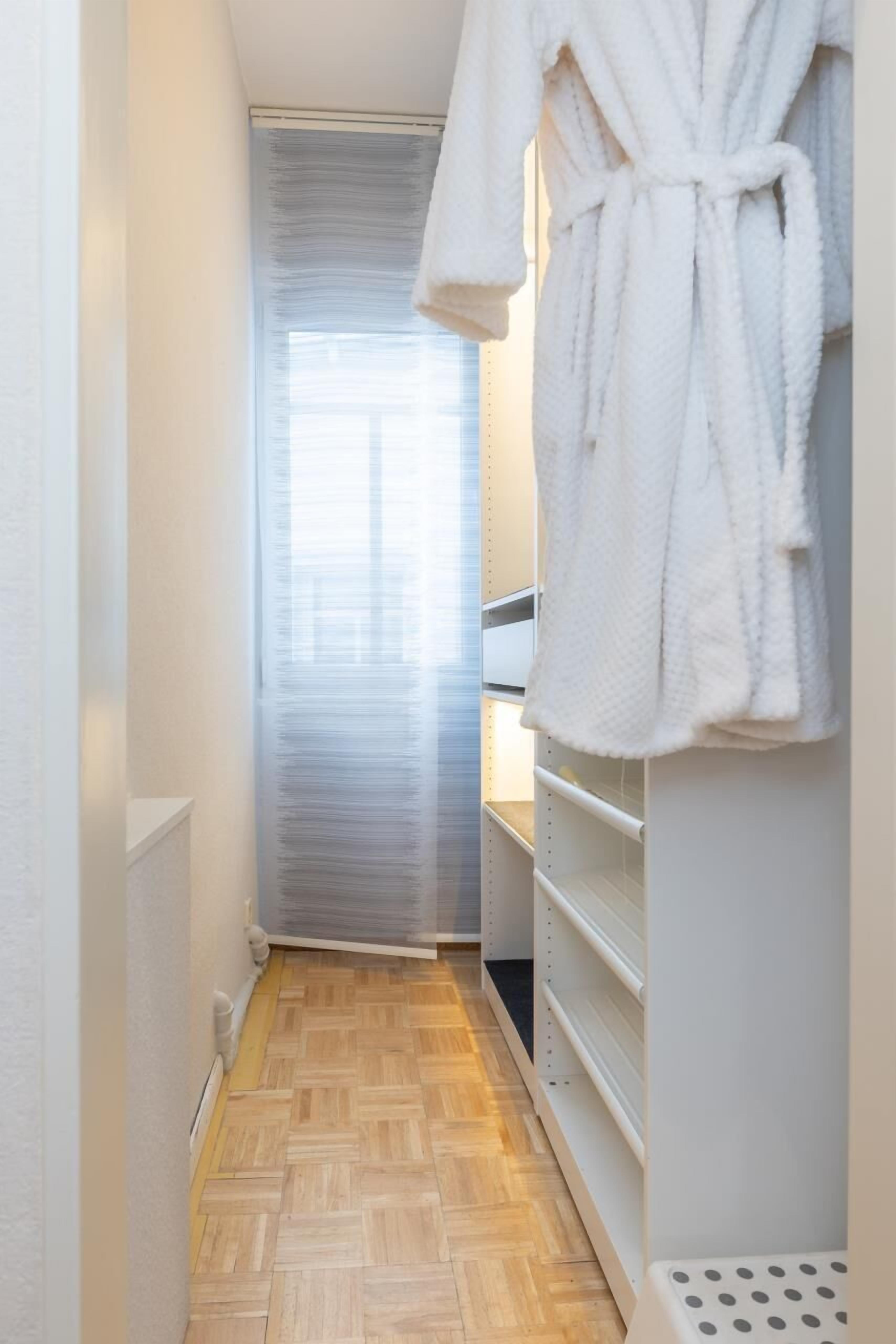 Um corredor estreito com piso de madeira, um roupão branco pendurado à direita e uma porta no final.
