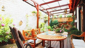 Een houten terras met een ronde tafel en stoelen, omringd door plantenpotten en een bakstenen muur.