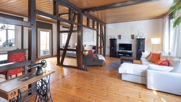 Een ruime woonkamer met houten vloeren, een grote houten balkenconstructie, een witte bank, een televisie en een naaimachine.