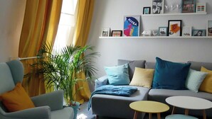 A living room with a grey sofa, a blue armchair, and a yellow table. There are plants, a bookshelf with framed pictures, and a window with yellow curtains.