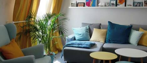 Ein Wohnzimmer mit einem grauen Sofa, einem blauen Sessel und einem gelben Tisch. Es gibt Pflanzen, eine Bücherwand mit gerahmten Bildern und ein Fenster mit gelben Vorhängen.