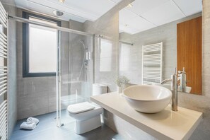 A modern bathroom with a glass shower enclosure, a white toilet, a large vessel sink, and a wooden cabinet. - Aspasios Diagonal Art (Barcelona)
