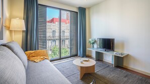 A modern living room with a sofa, a coffee table, a TV on a stand, and a view of buildings through large windows. - Aspasios Diagonal Art (Barcelona)