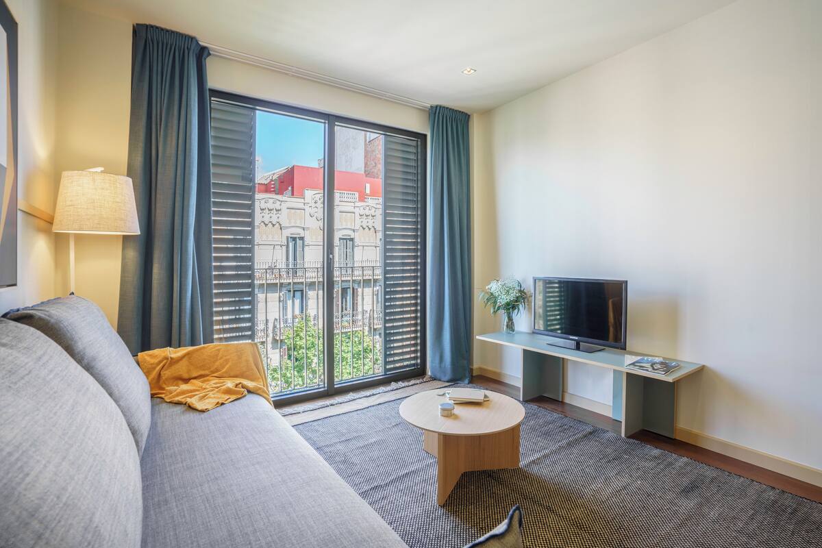 A modern living room with a sofa, a coffee table, a TV on a stand, and a view of buildings through large windows.