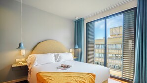 A hotel room with a large bed, a nightstand, a lamp, and a view of the city through the window. - Aspasios Diagonal Art (Barcelona)
