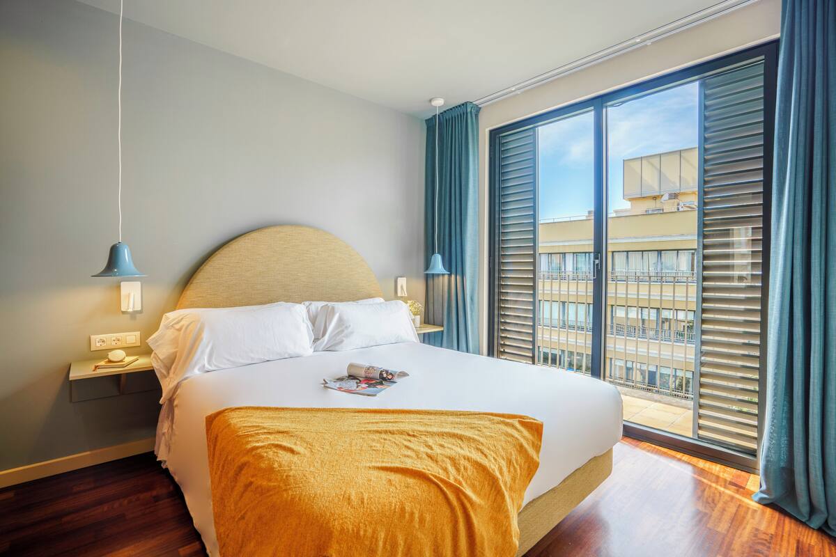 A hotel room with a large bed, a nightstand, a lamp, and a view of the city through the window.