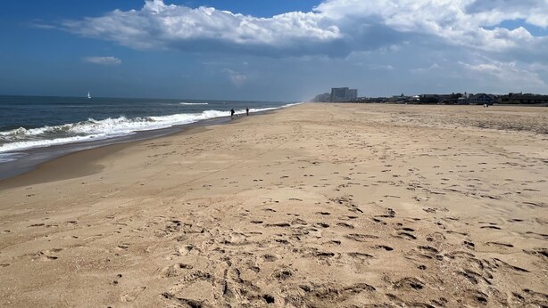 Uma extensa praia de areia com pegadas, um veleiro ao longe e edifícios ao longo da costa.