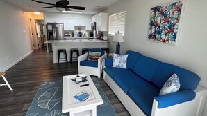 Uma sala de estar moderna com um sofá azul, poltronas brancas e uma mesa de centro. Uma cozinha com armários brancos e um espaço de bar é visível ao fundo.