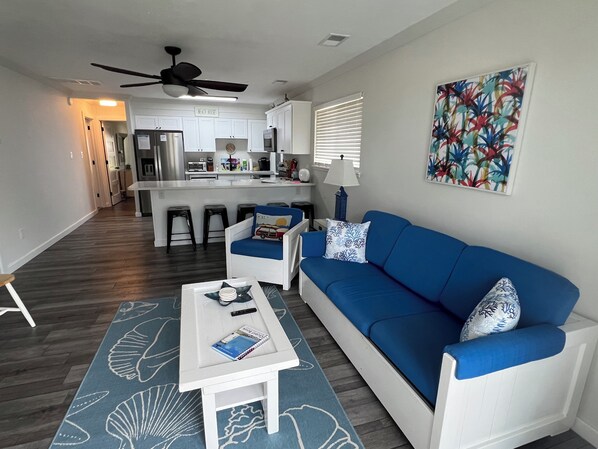 A modern living room with a blue sofa, white armchairs, and a coffee table. A kitchen with white cabinets and a bar area is visible in the background.