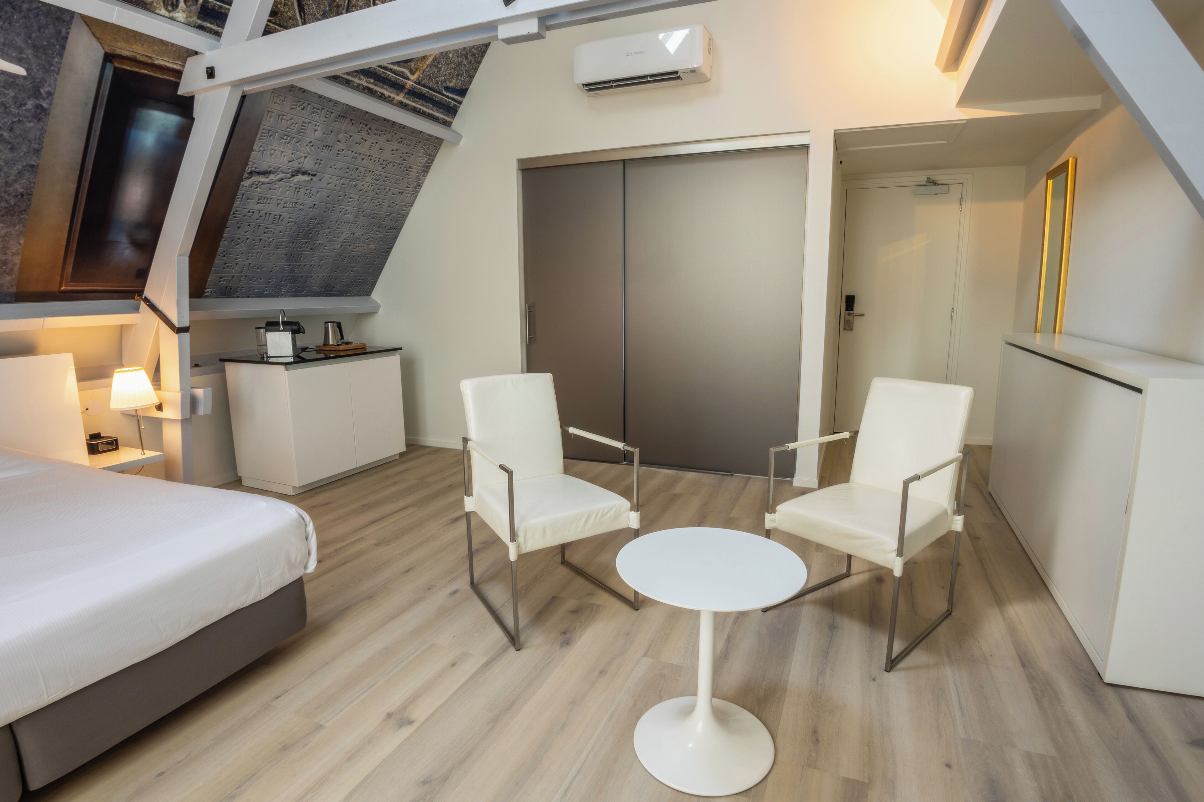 A modern hotel room with a bed, two white chairs, a small round table, a kitchenette, and a large wardrobe.