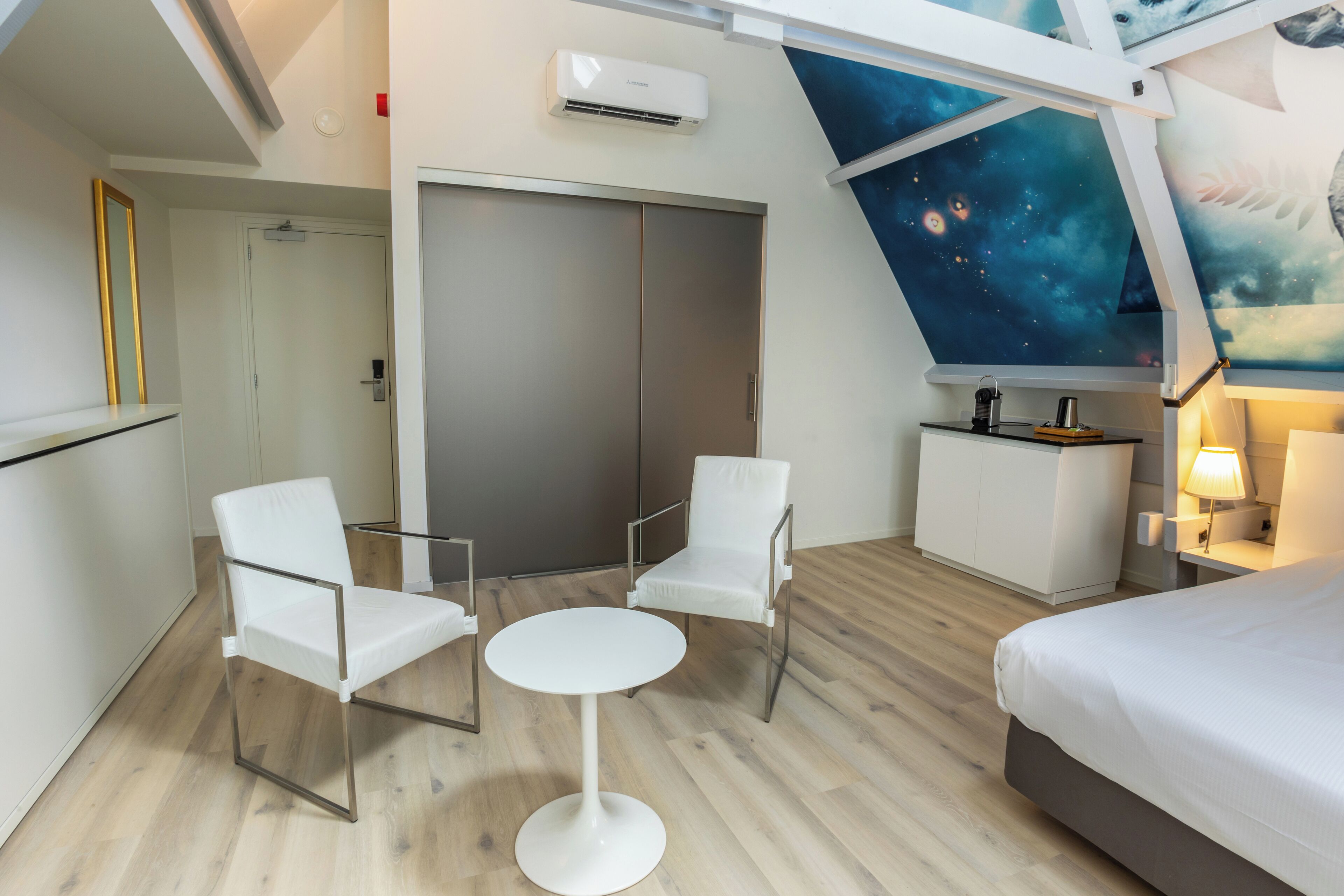 Een moderne hotelkamer met een bed, twee witte stoelen, een klein rond tafeltje, een bureau met stoel, een garderobe en een plafond waarop een muurschildering van een sterrennacht is afgebeeld.