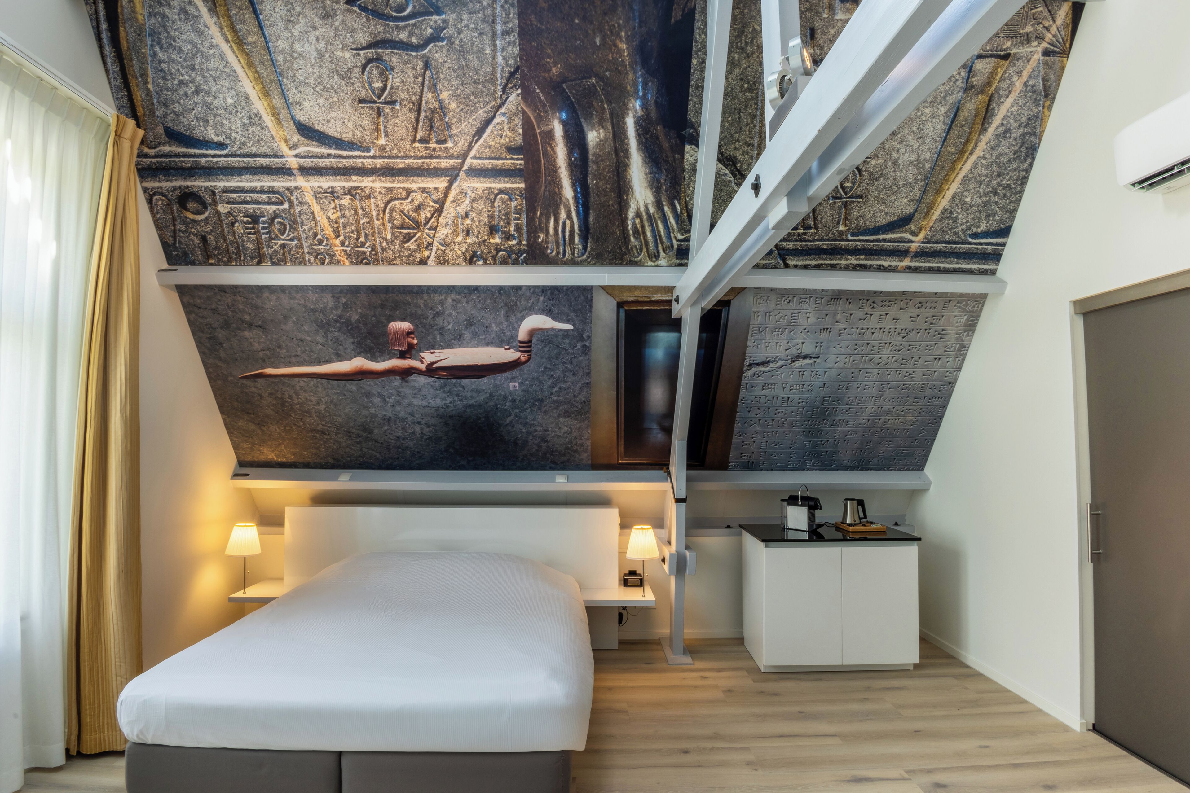 Una camera d'albergo moderna con un letto grande, una scrivania e un affresco raffigurante geroglifici egizi antichi.