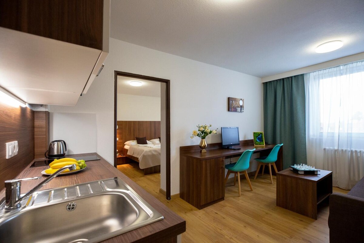 A compact hotel room with a kitchenette, a bed, a desk with a computer, two chairs, and a large window with curtains.