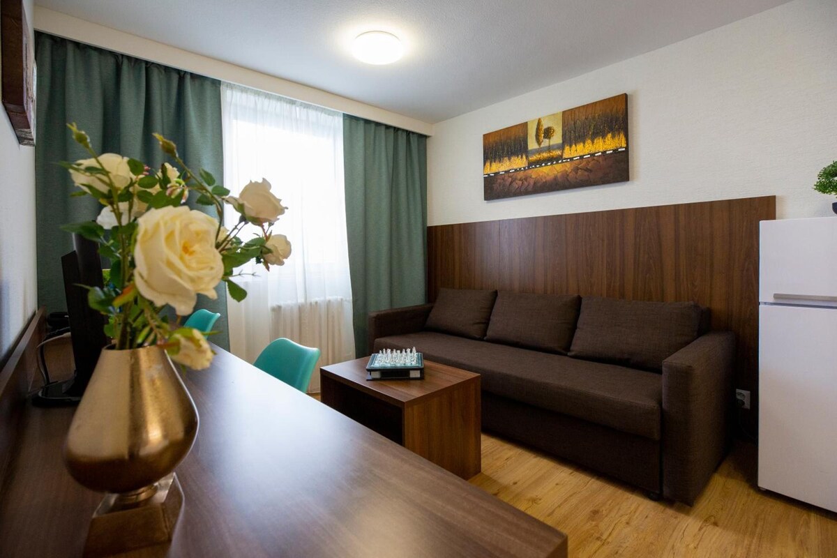 A modern living room with a wooden sofa, a coffee table, and a chessboard. There is a painting on the wall and a vase with flowers on the table.
