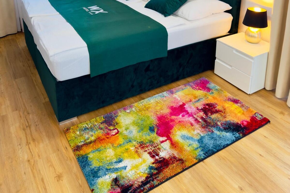 A neatly made bed with a green bedspread and matching pillows, a colorful rug, a bedside table with a lamp, and a painting of a tree on the wall.