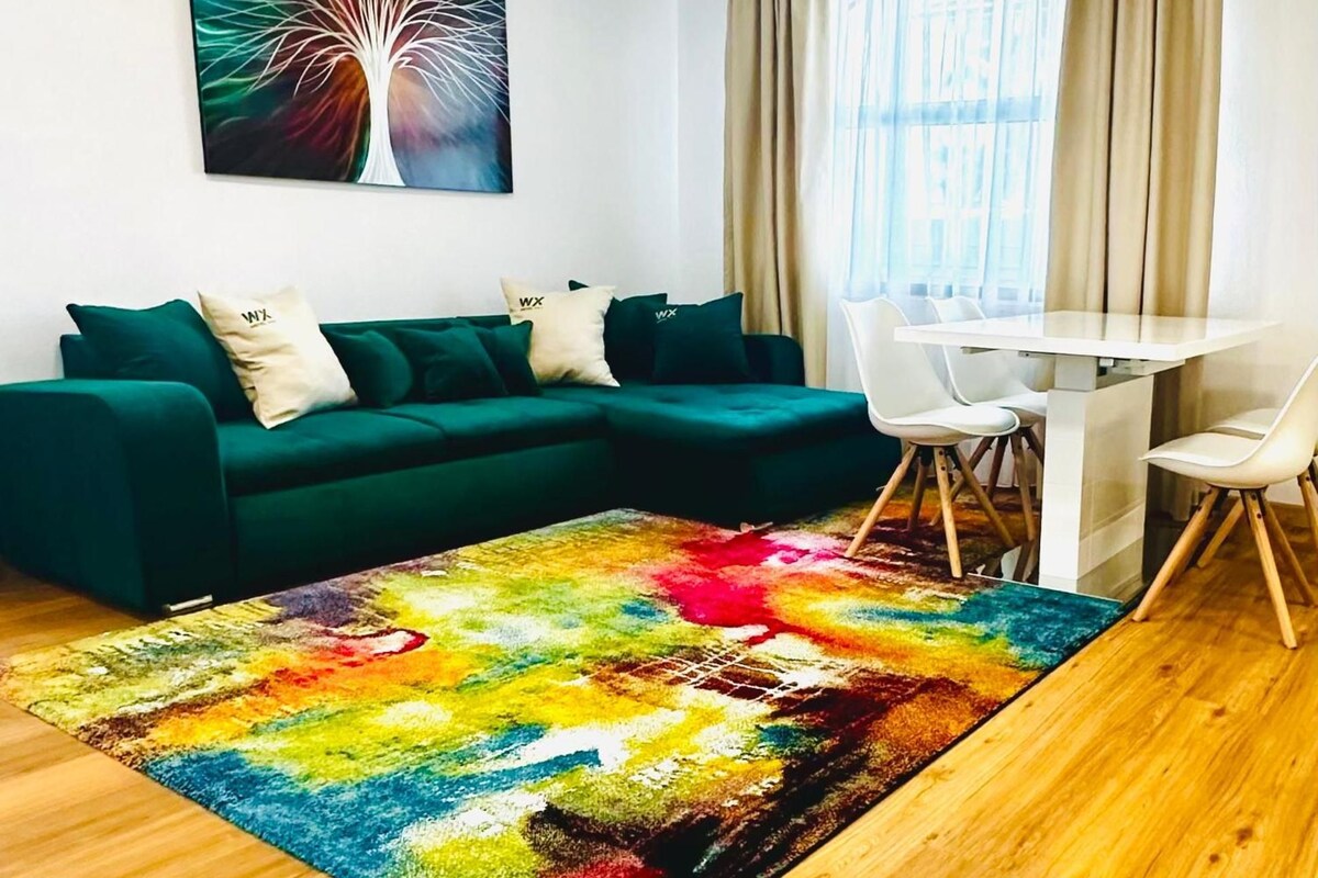 Un salon avec un canapé vert, un tapis coloré, une table à manger avec des chaises et une fenêtre ornée de rideaux beiges.