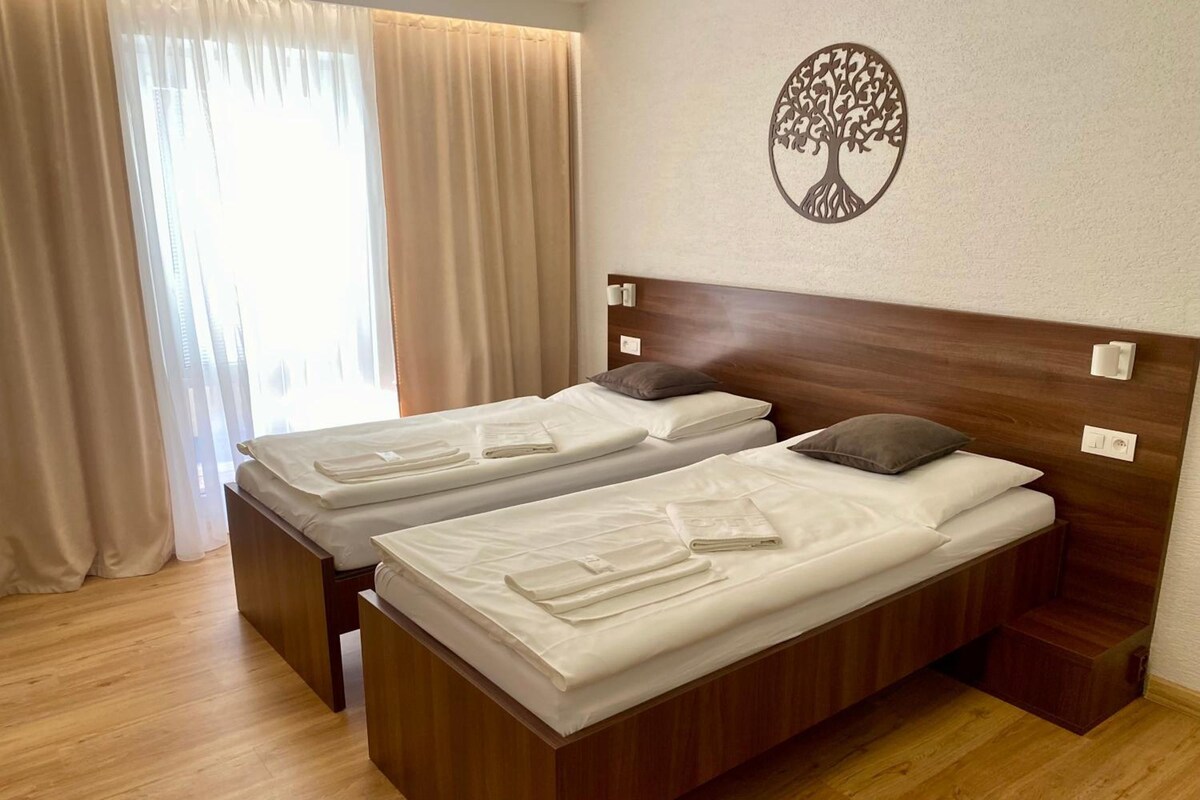 A hotel room with two beds, wooden headboards, a wall-mounted tree decoration, and a window with curtains.