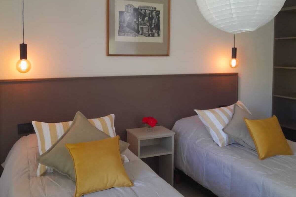 Habitación de hotel con dos camas, cada una con un almohadón y una mesita de noche. Hay un cuadro en la pared y un farol de papel blanco colgando del techo.