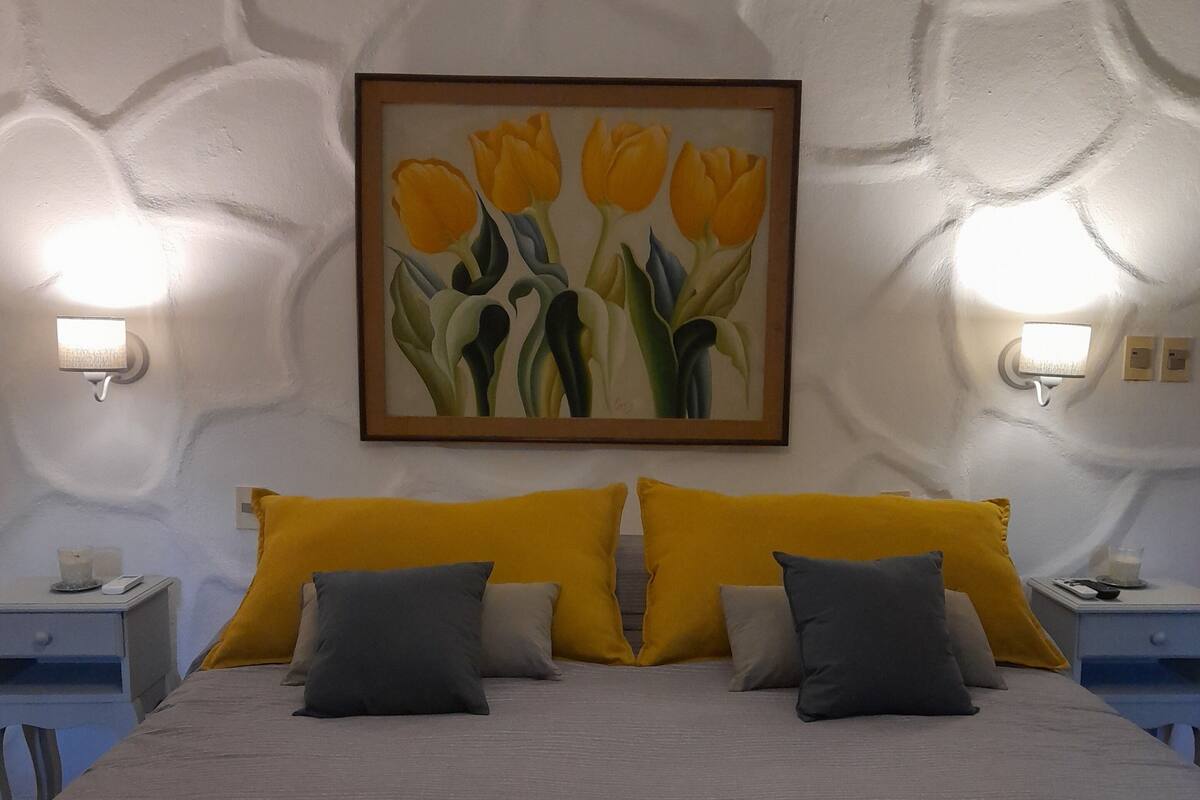 Una cama con colcha estampada, dos mesitas de noche, un cuadro colgado en la pared con tulipanes amarillos y dos lámparas de pared.