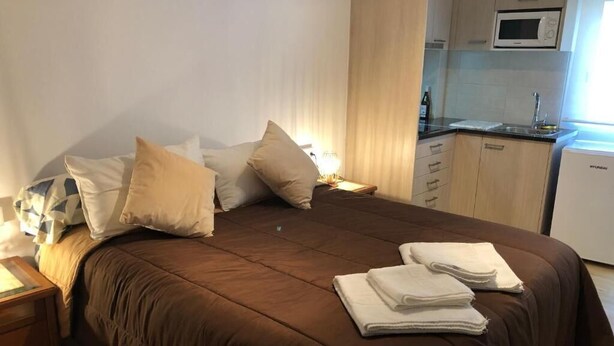 Una cama bien hecha con ropa de cama marrón, una mesita de noche con lámpara, y una zona de cocina con microondas y fregadero.
