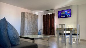 Una sala moderna con sofá, una mesa de centro de vidrio, zona de comedor con sillas, televisor montado en la pared y un revestimiento de pared de madera.