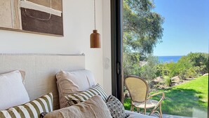 Un dormitorio con una cama grande, varios almohadones, una lámpara colgante y vistas a un jardín exuberante y al océano a través de un ventanal.