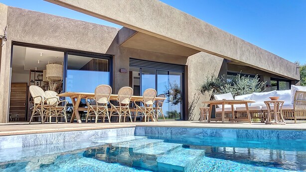 Un área de piscina moderna con agua azul cristalina, mobiliario de mimbre y un edificio contemporáneo con grandes ventanales.