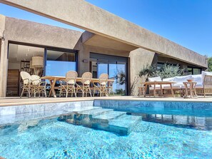 A modern pool area with clear blue water, wicker furniture, and a contemporary building with large windows. - Superbe Villa vue Mer, Piscine Chauffée, Terrain de Pétanque (Corbara)